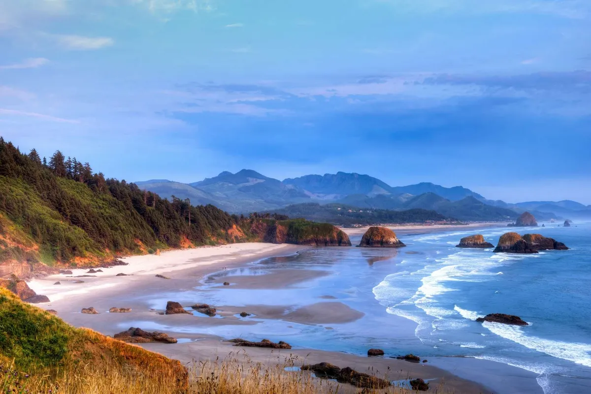 A panoramic view of the Oregon coastline with sandy beaches, ocean waves, dramatic rock formations, and distant mountains. The natural beauty and expansive scenery invite exploration and day trips along the coast.