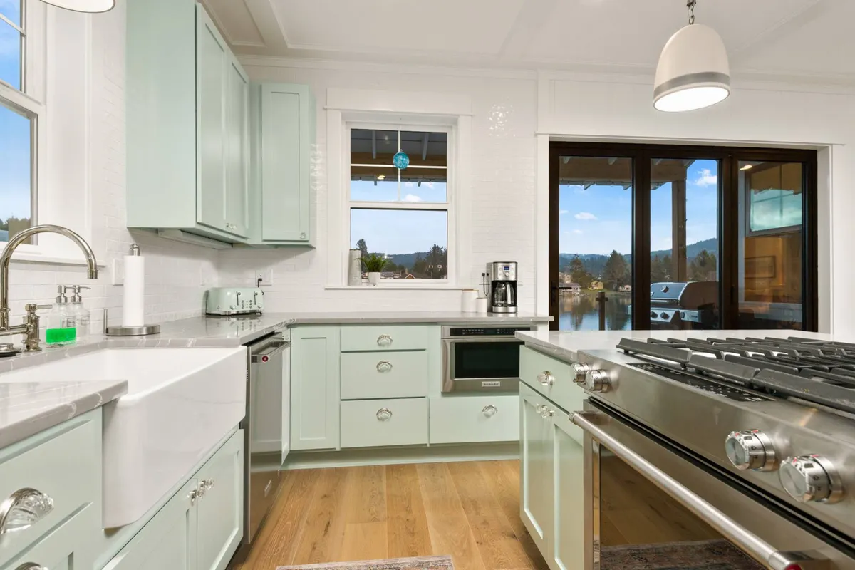A spacious, open chef’s kitchen with mint green cabinetry and stainless steel appliances. Large windows bathe the space in natural light, offering scenic views while allowing guests to gather around the central island for cooking and conversation. The kitchen seamlessly connects to adjacent living areas, creating a sense of togetherness.