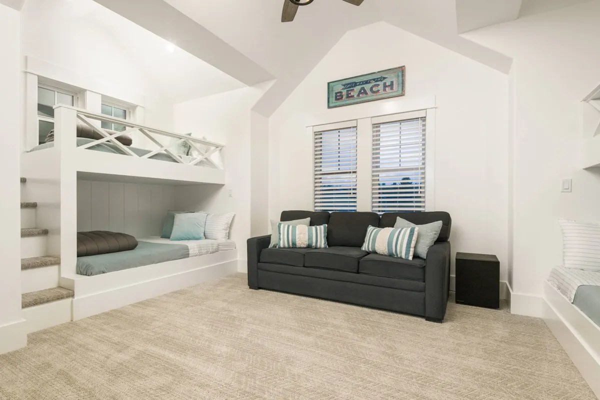 The inviting bunk/media room is shown with a sturdy bunk bed, a comfortable sofa, window blinds letting in natural light, and a beach-themed decorative sign. The space exudes playfulness and comfort, ideal for kids or group hangouts.