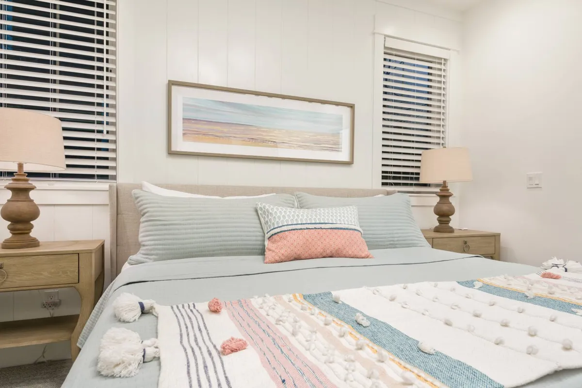 A queen bedroom is depicted with serene décor, decorative pillows, a nightstand with lamp, and framed artwork. The room glows with natural light and offers a peaceful atmosphere for relaxation or a restful night's sleep.
