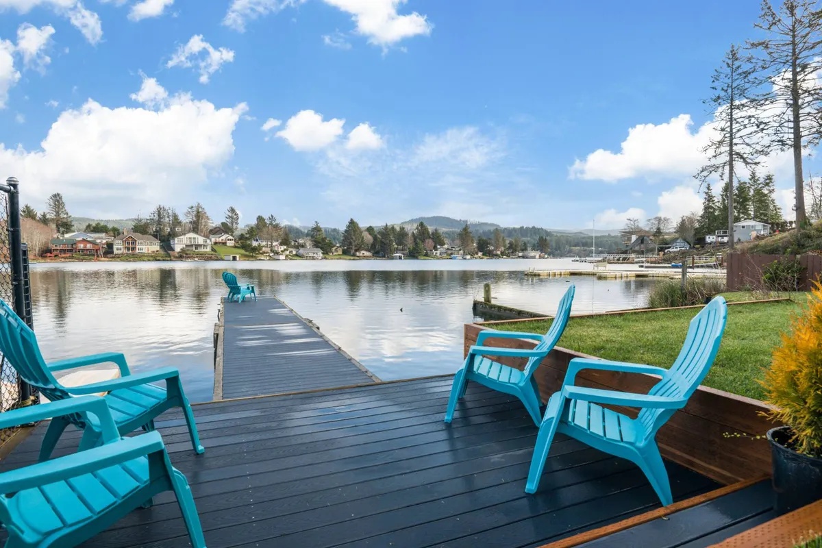 A serene view from the dock, featuring Adirondack chairs positioned for relaxing, with sweeping views over the lake and surrounding natural landscape. The calm water and seating suggest tranquil mornings or evenings by the lake.