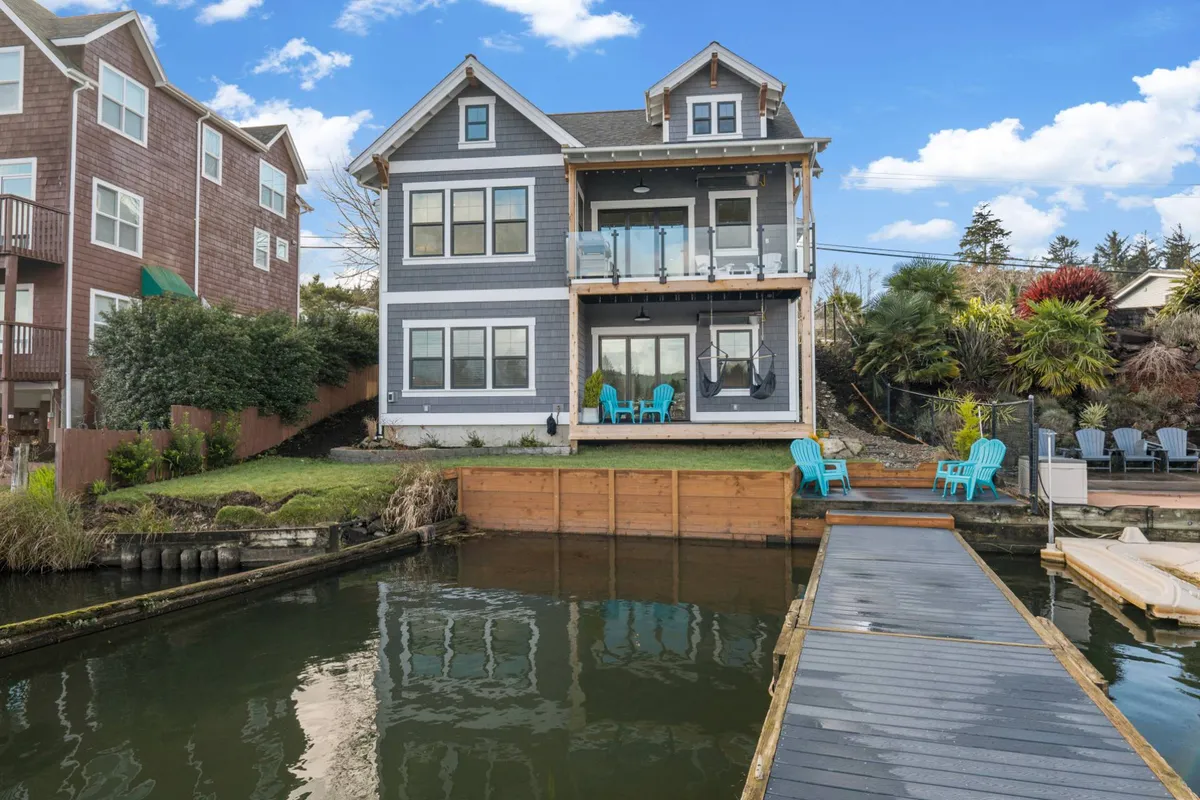 A wide exterior shot of the property, featuring a modern deck with blue chairs and a dock stretching into the lake. The scene encompasses both the lounging deck and direct water access, capturing the transition from comfort to adventure.