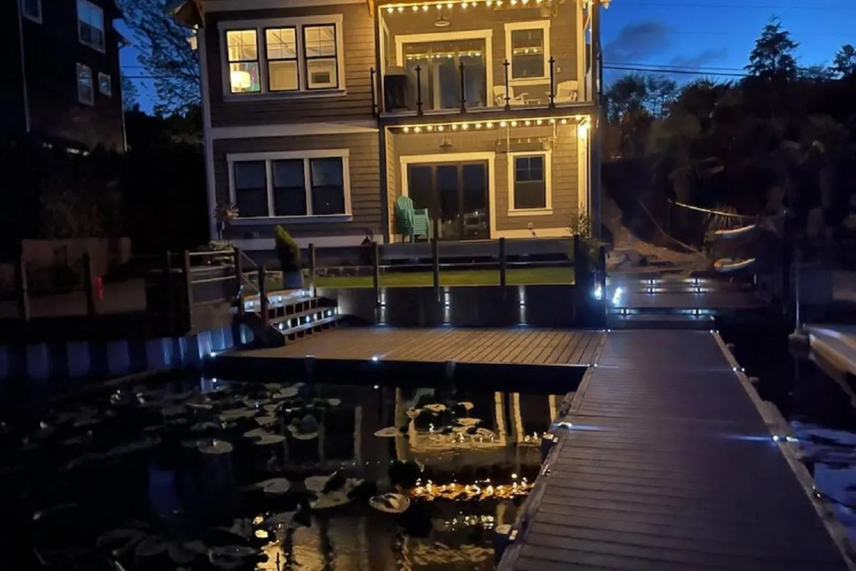This image features the illuminated house at dusk, a wooden dock jutting over calm water adorned with lily pads, and an inviting patio where evening gatherings can take place. The tranquil, cozy mood is enhanced by the warm lighting and serene setting.