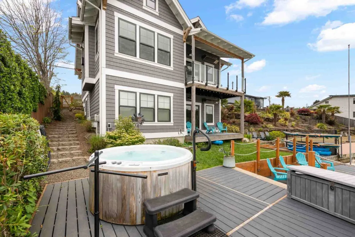 An exterior view featuring a hot tub on the deck, outdoor seating, landscaped garden, and the house in the background. The hot tub is highlighted as the perfect spot for stargazing or soaking after a fun-filled day.