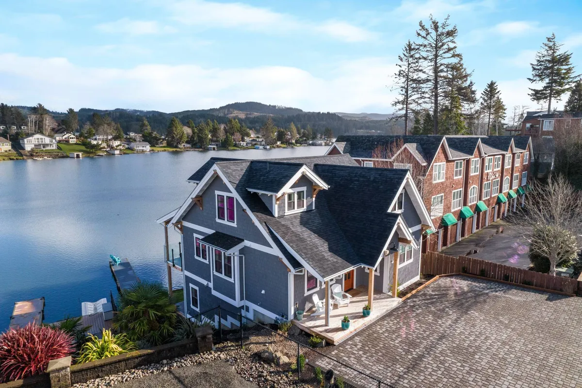 The image showcases a stunning, custom-built lakefront home set against lush trees, with the tranquil lake stretching out in front. The golden glow of sunset reflects off the water, highlighting the inviting porch perfect for relaxing or taking in the view. In the background, hints of nearby recreational activities—like a dock for boating and areas for picnicking—suggest the many adventures awaiting guests.